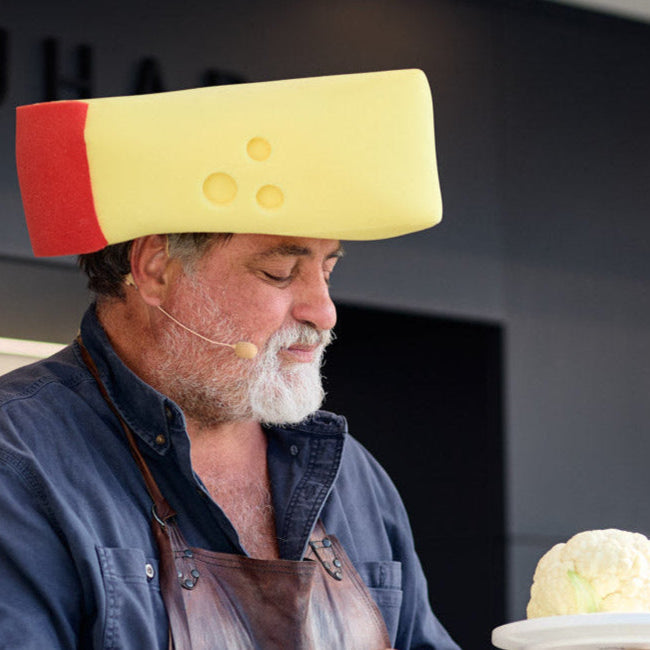 Person wearing a large cheese head with a cauliflower on a plate in front of them, with 'QUA' visible in the background.